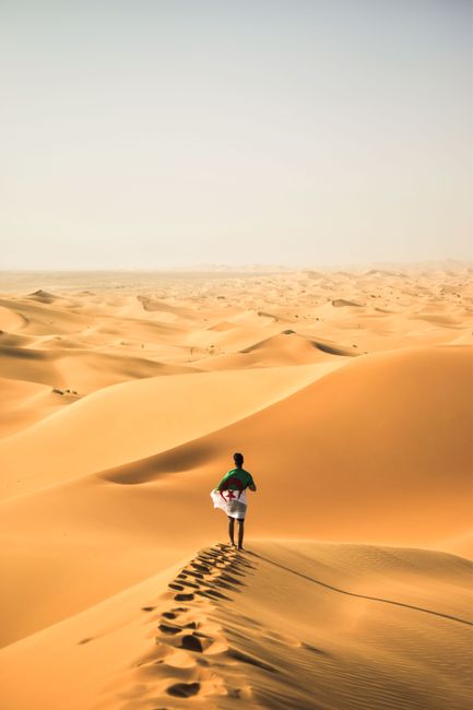 A solitary traveler walks across the vast sand dunes of the Sahara Desert in Algeria.
