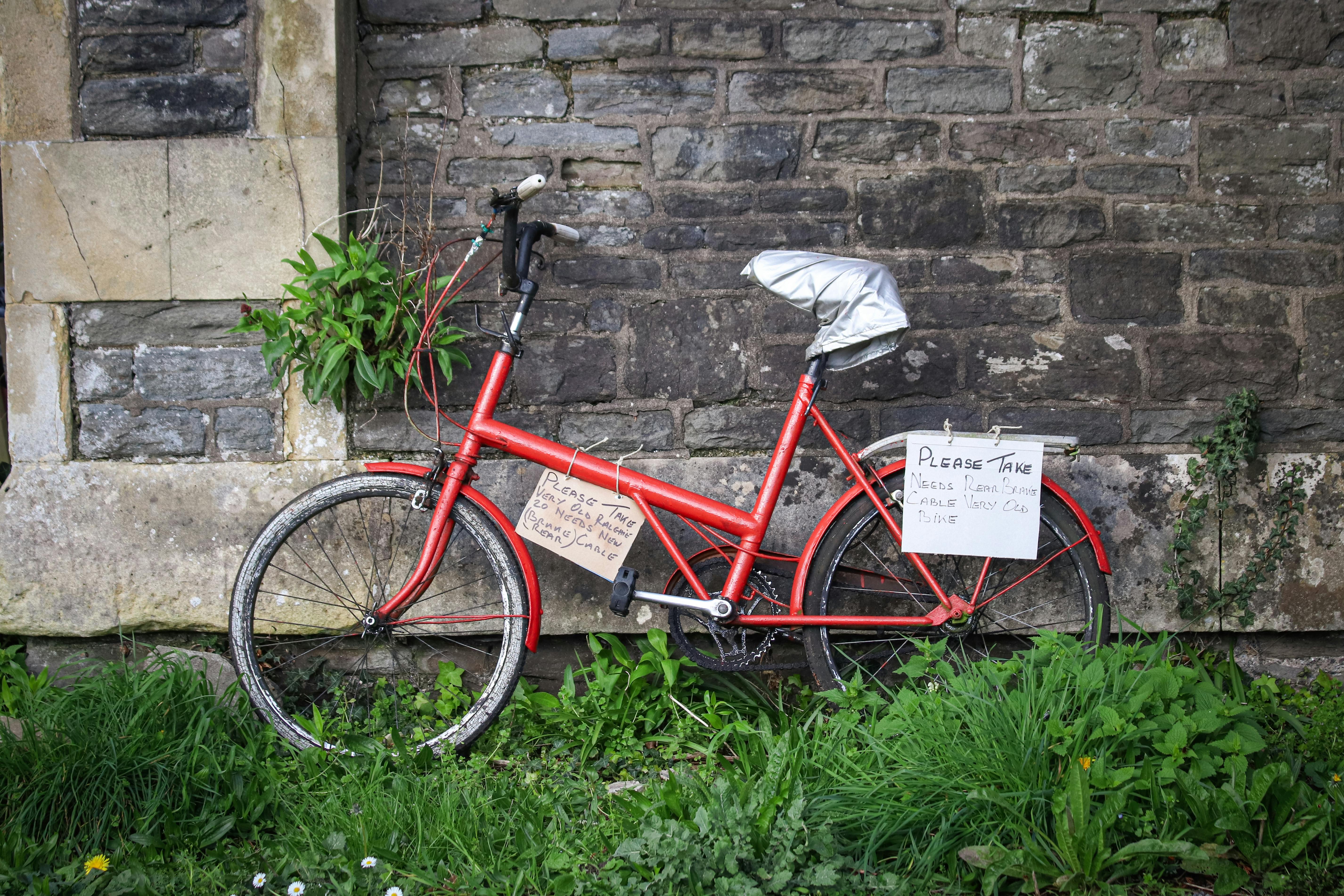 Free A rusty red bicycle leaning against a stone wall with foliage and signs. Stock Photo