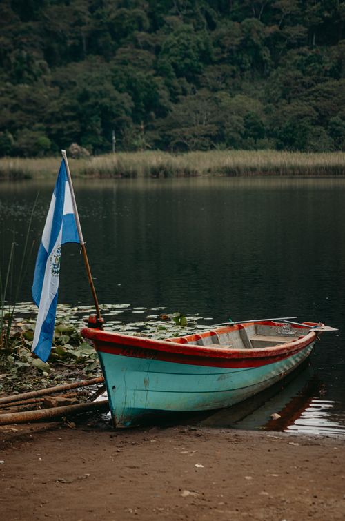 Free A vibrant canoe with a flag by a tranquil lake, surrounded by lush forest. Stock Photo