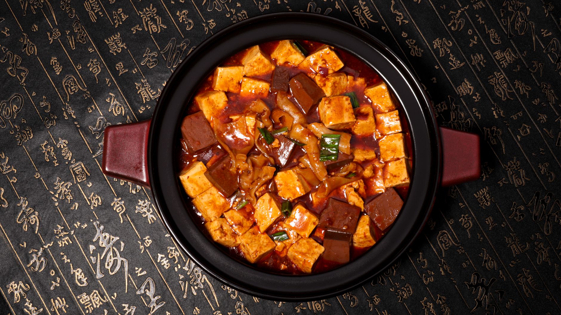 Cubes of silky white tofu swimming in glossy, dark red mapo sauce garnished with bright green scallions in a white ceramic bowl