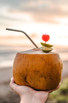 Hand holding coconut with straw and fruit garnish at sunset by the beach.
