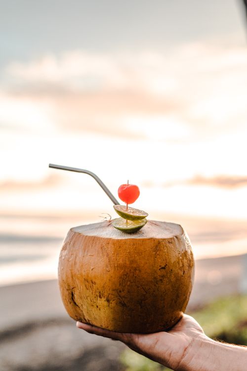 Free Exotic coconut water with a cherry and lime garnish at a beach sunset, representing tropical freshness. Stock Photo