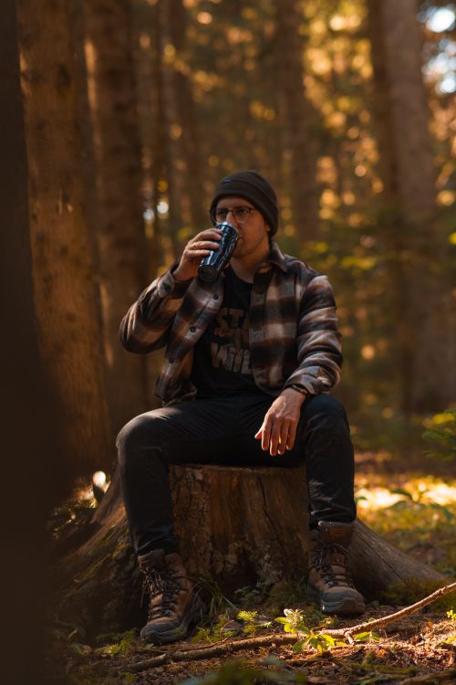 Free Man sits on a tree trunk in the forest, sipping from a thermos, evoking a tranquil outdoor experience. Stock Photo