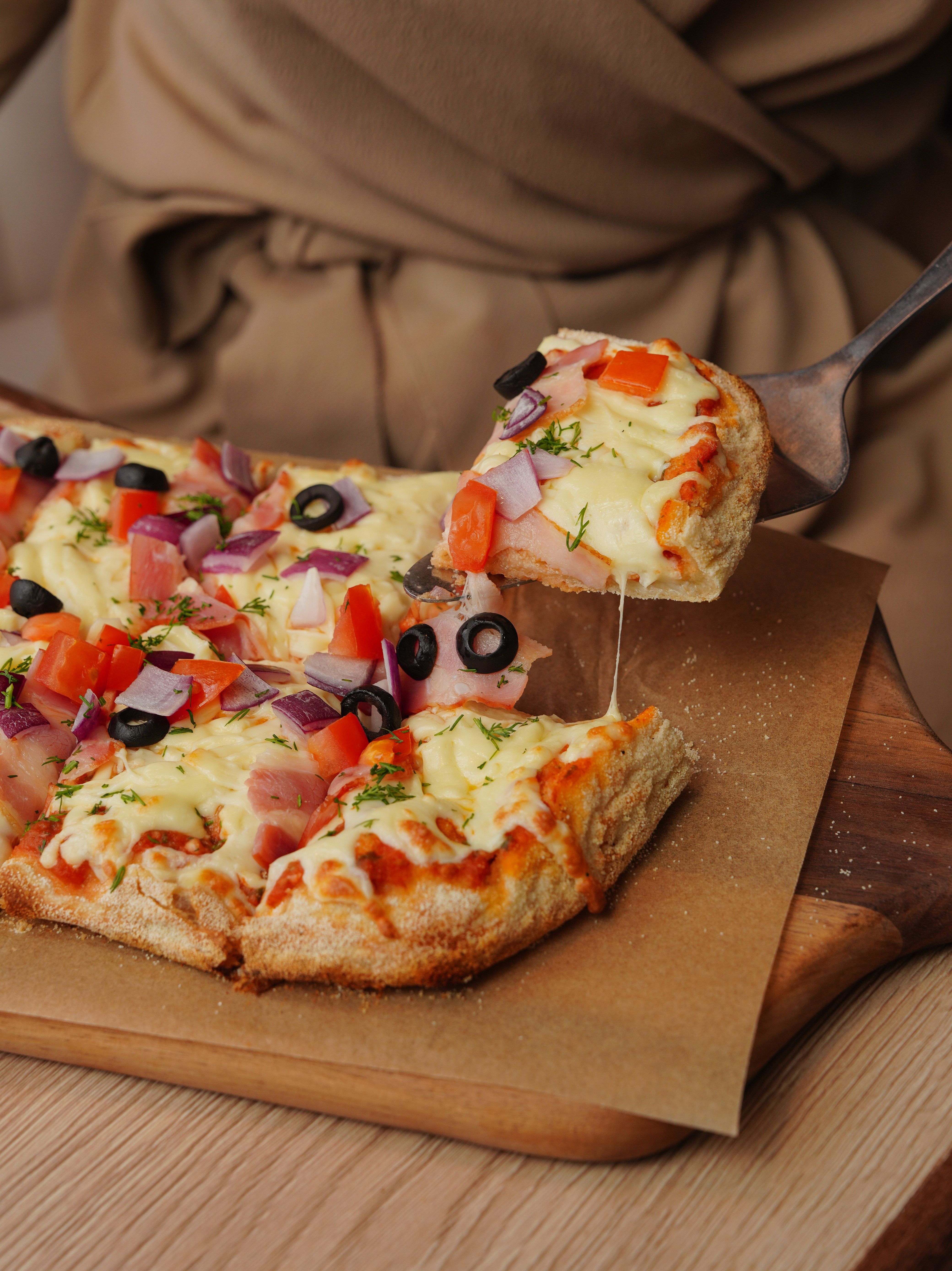 Appetizing vegetable pizza slice with toppings of olives, tomatoes, and cheese being lifted from a wooden board.