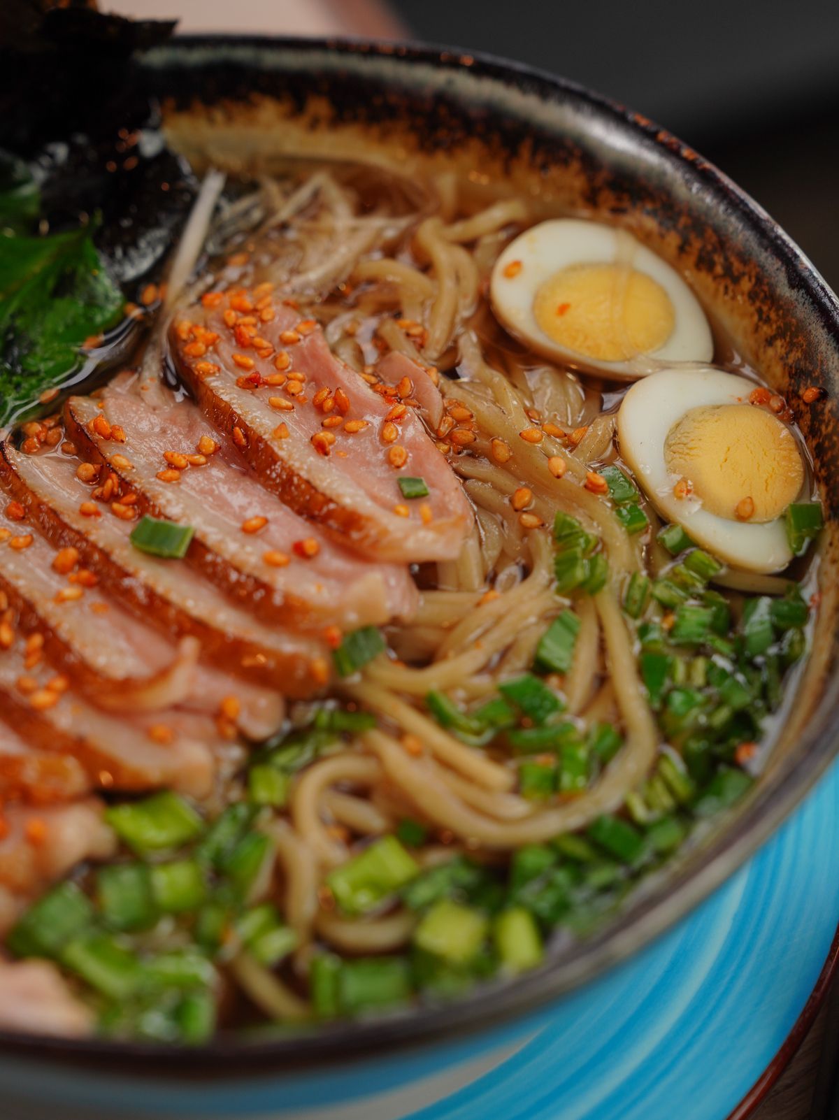 Close-up of beef ramen bowl with pink slices of beef, halved boiled eggs, chopped scallions, noodles and toasted sesame seeds in dark broth
