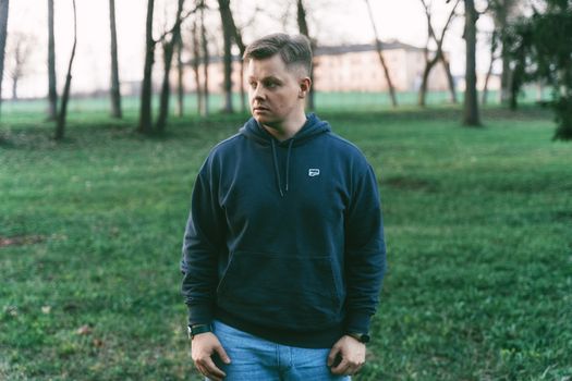 A man in a hoodie stands thoughtfully in a grassy woodland area, looking away from the camera.