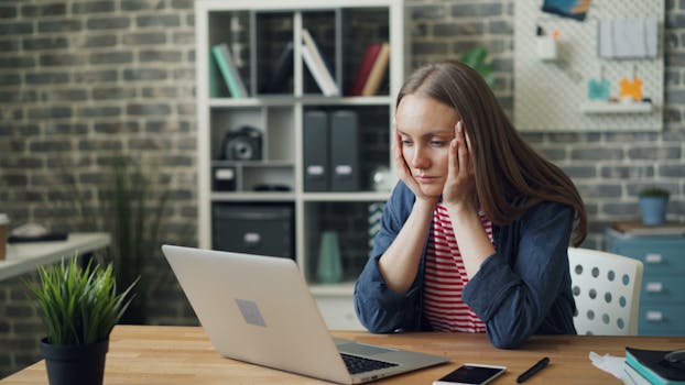 A woman appears stressed while working on a laptop in her modern office.