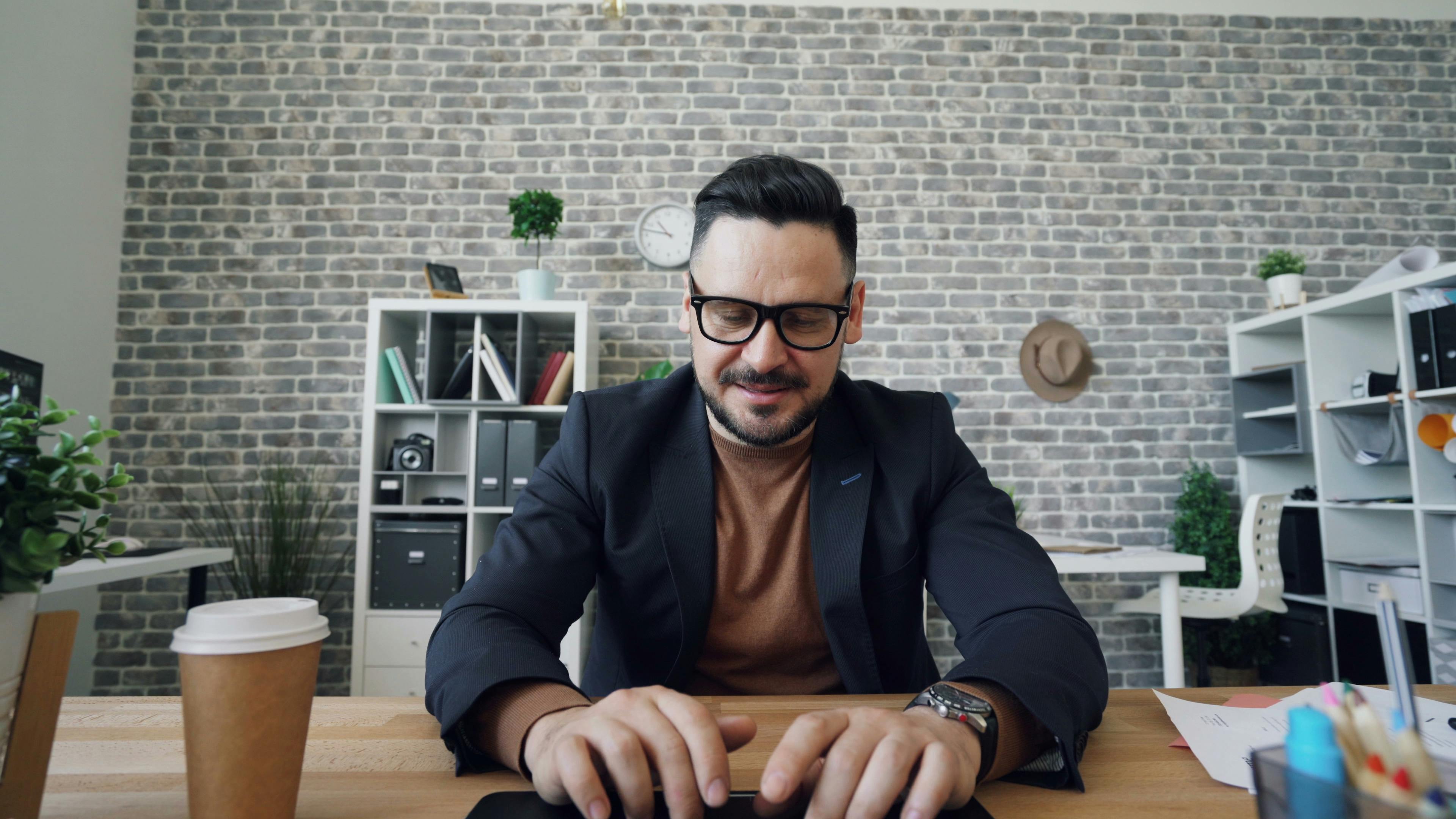 A man in glasses smiles while typing on a laptop in a stylish office.
