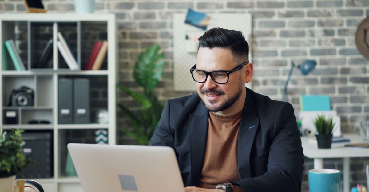 A person smiling while typing on a laptop with a coffee cup and a plant in a cozy home office