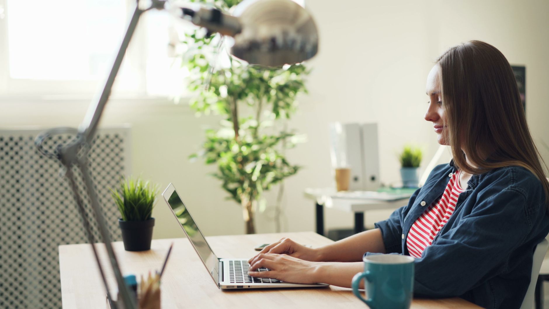 Woman smiling while working on a laptop in a modern office with plants.