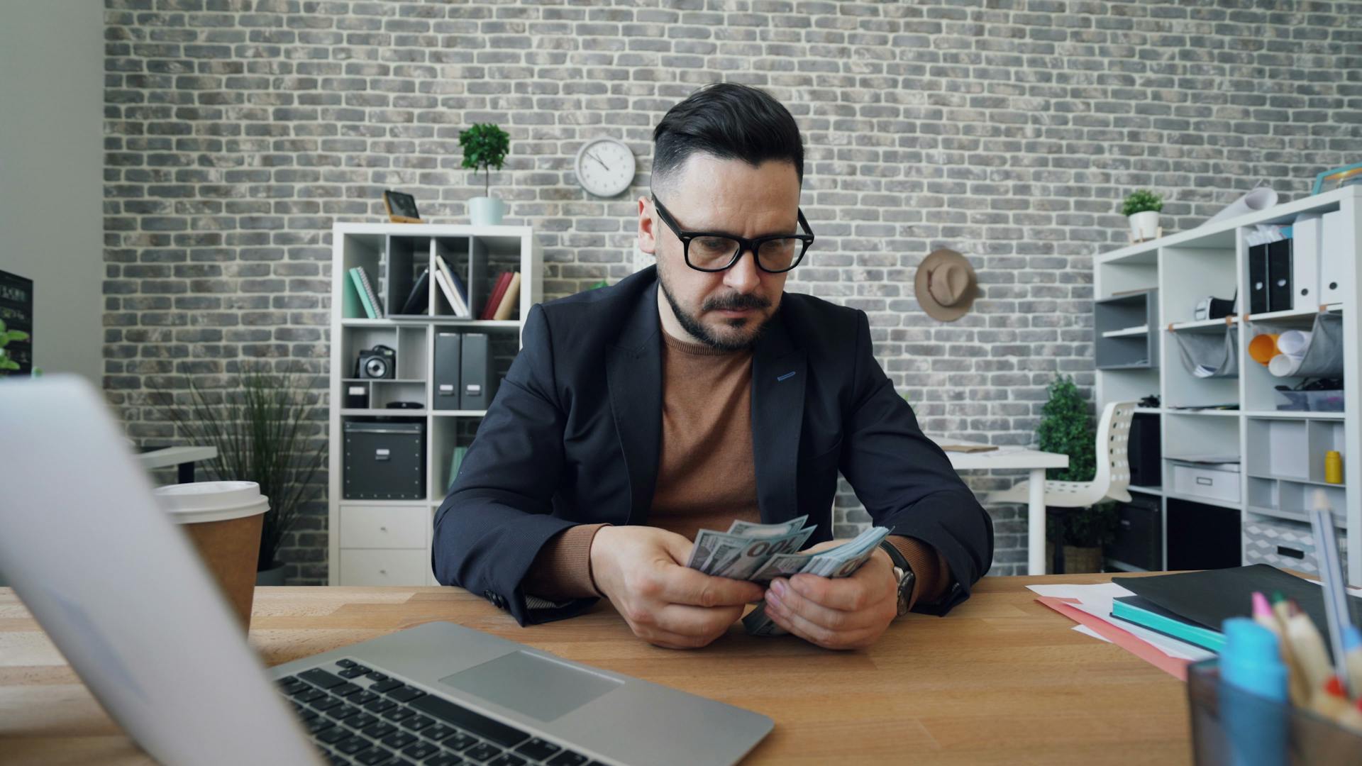 Businessman counting cash at a desk in a modern office setting with a laptop and documents.