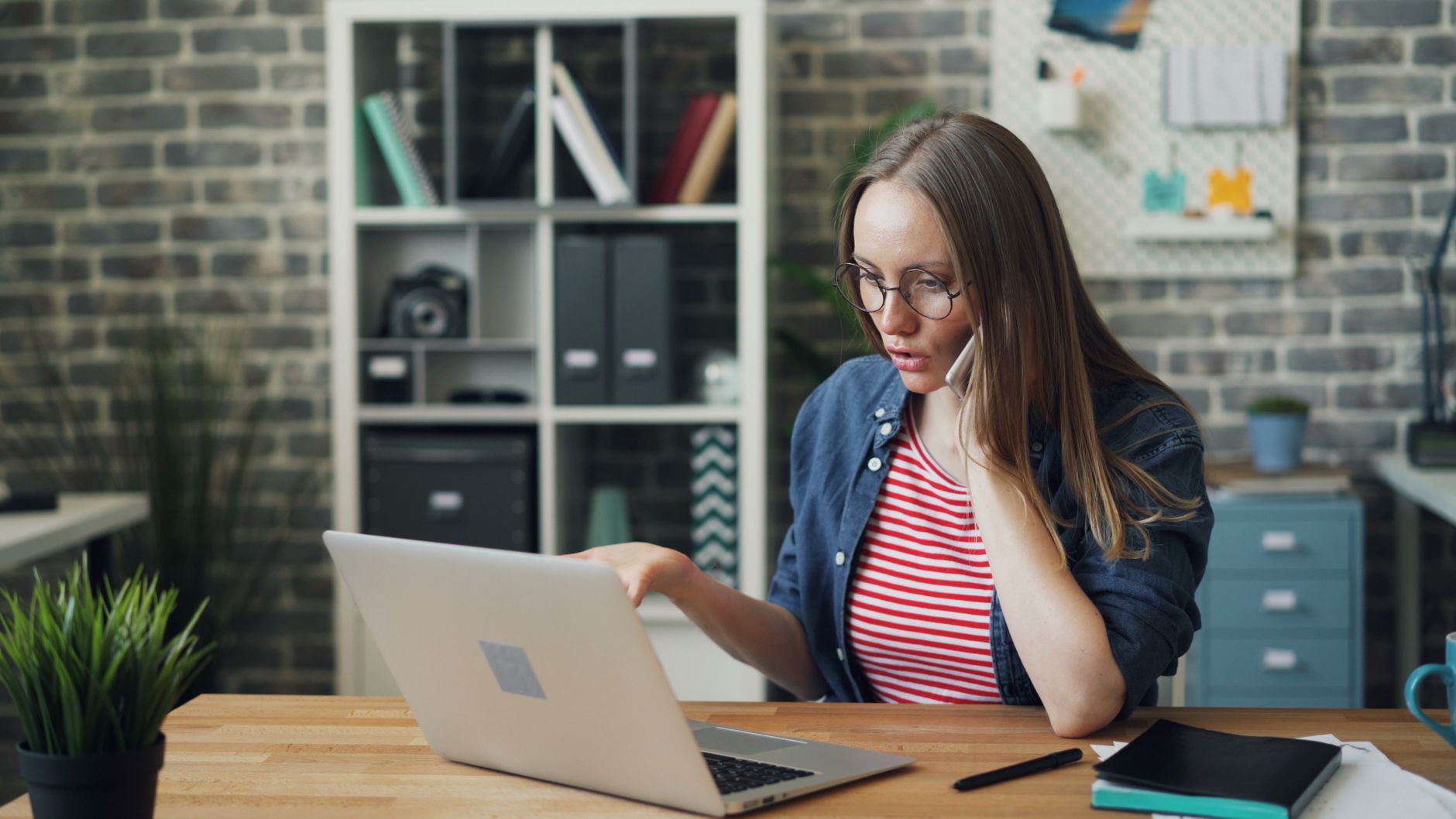 Focused young woman working on laptop while talking on a phone in a stylish modern office.