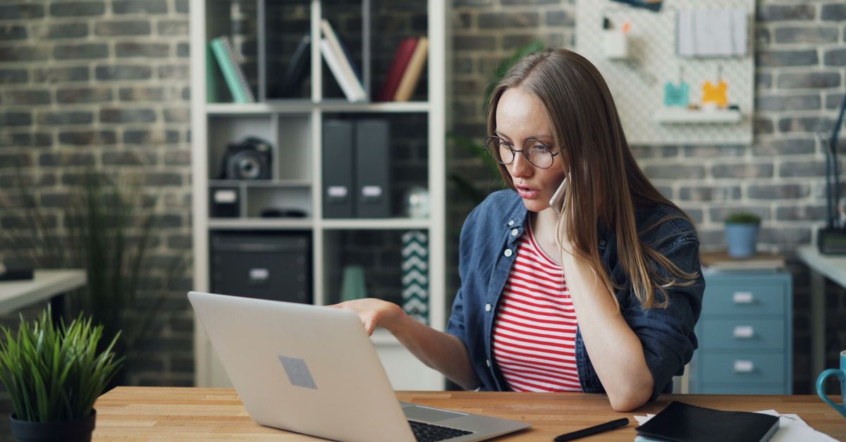 Focused young woman working on laptop while talking on a phone in a stylish modern office.