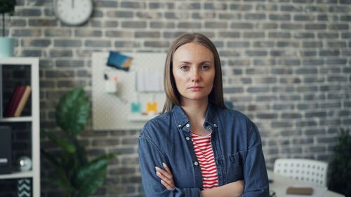 Free A confident woman stands with arms crossed in a modern office with a brick wall backdrop. Stock Photo