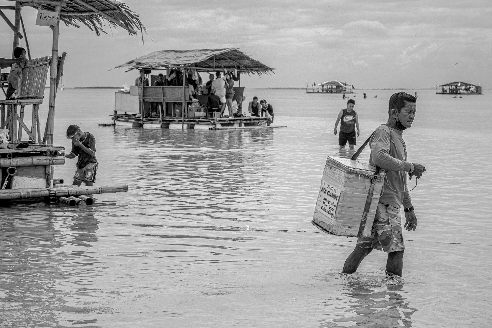 Black and white image of a man wading through the sea beside roofed rafts in a tropical setting.