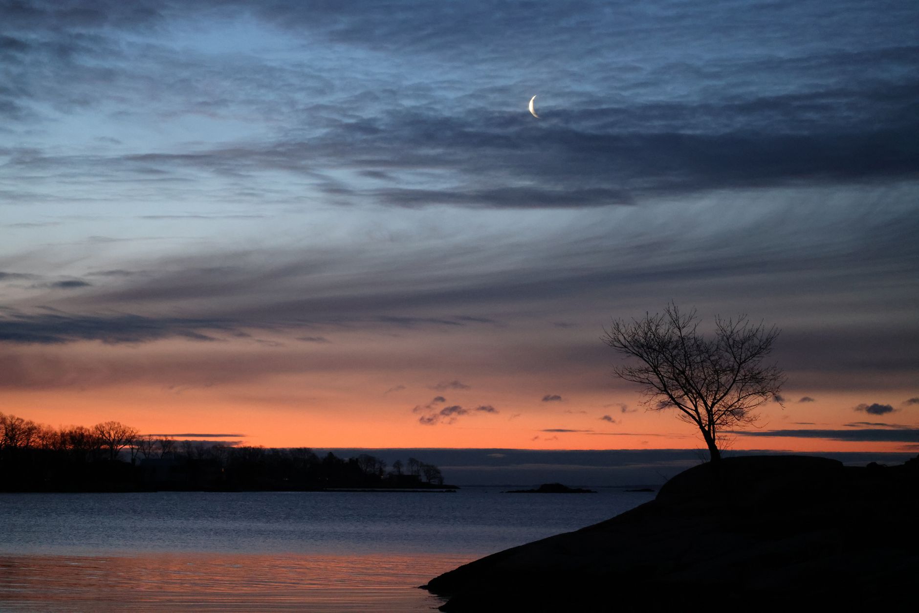 Tranquil lakeside view at sunset with silhouettes of trees and a crescent moon.