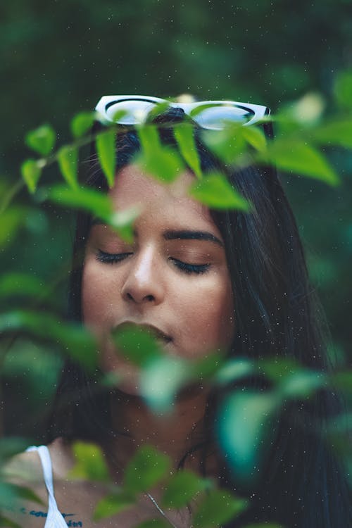 Фотография женщины, стоящей за зелеными листьями с закрытыми глазами