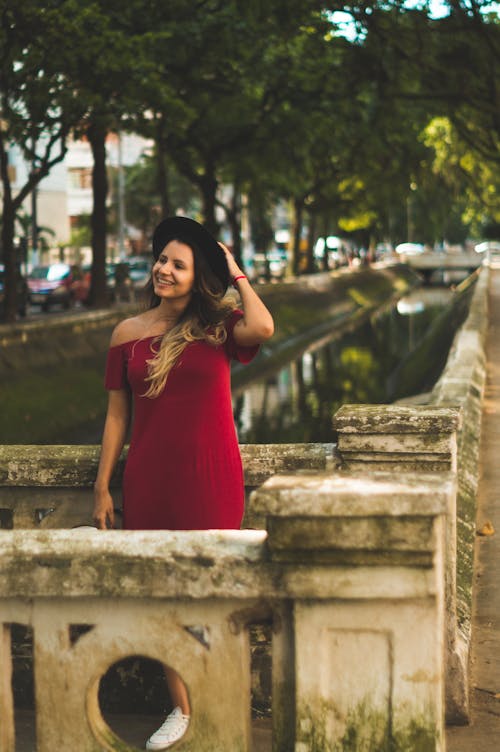Foto Der Lächelnden Frau Im Schwarzen Hut Und Im Roten Kleid, Die Auf Grauer Betonbrücke Stehen