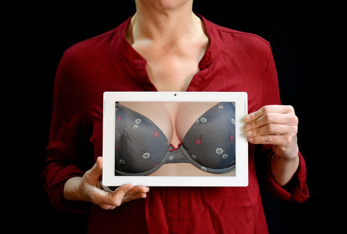 A person holding a tablet in front of their chest. The tablet displays breasts in a gray bra.