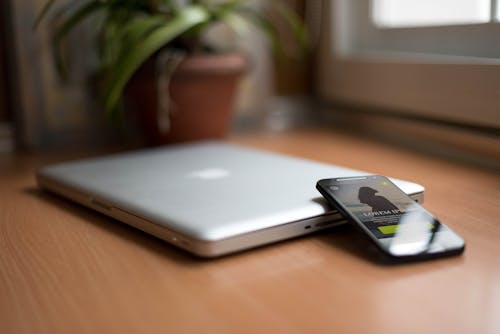 бесплатная смартфон рядом с серебряным Macbook на коричневом деревянном столе с горшечным растением на заднем плане Стоковое фото