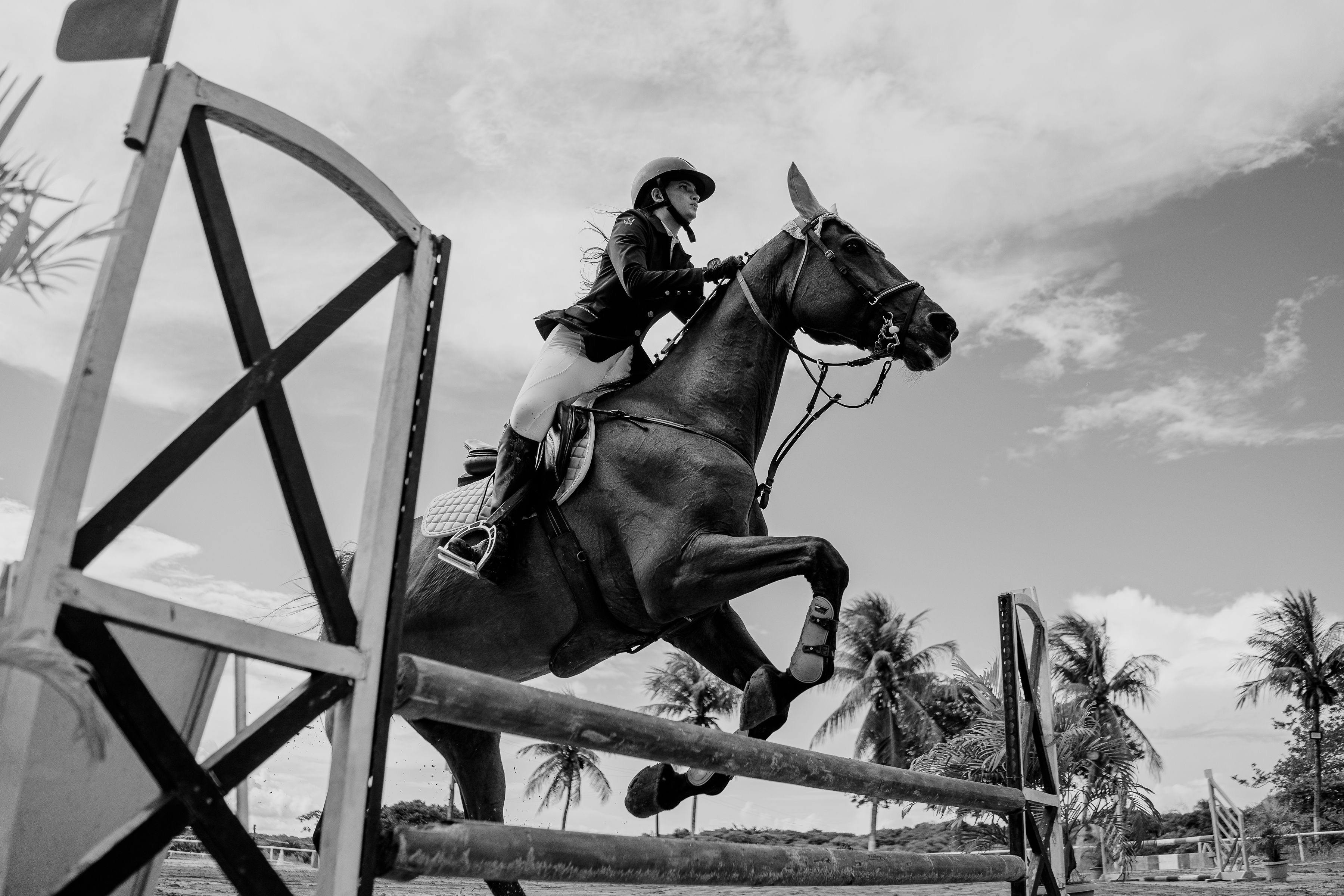 A woman riding a horse mid-jump during an equestrian event in a tropical outdoor setting.