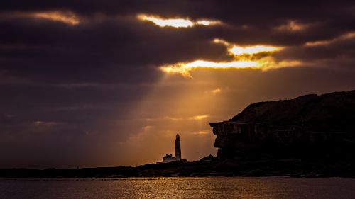 Free Silhouette of a lighthouse against a dramatic sunset sky on a rocky coastline. Stock Photo