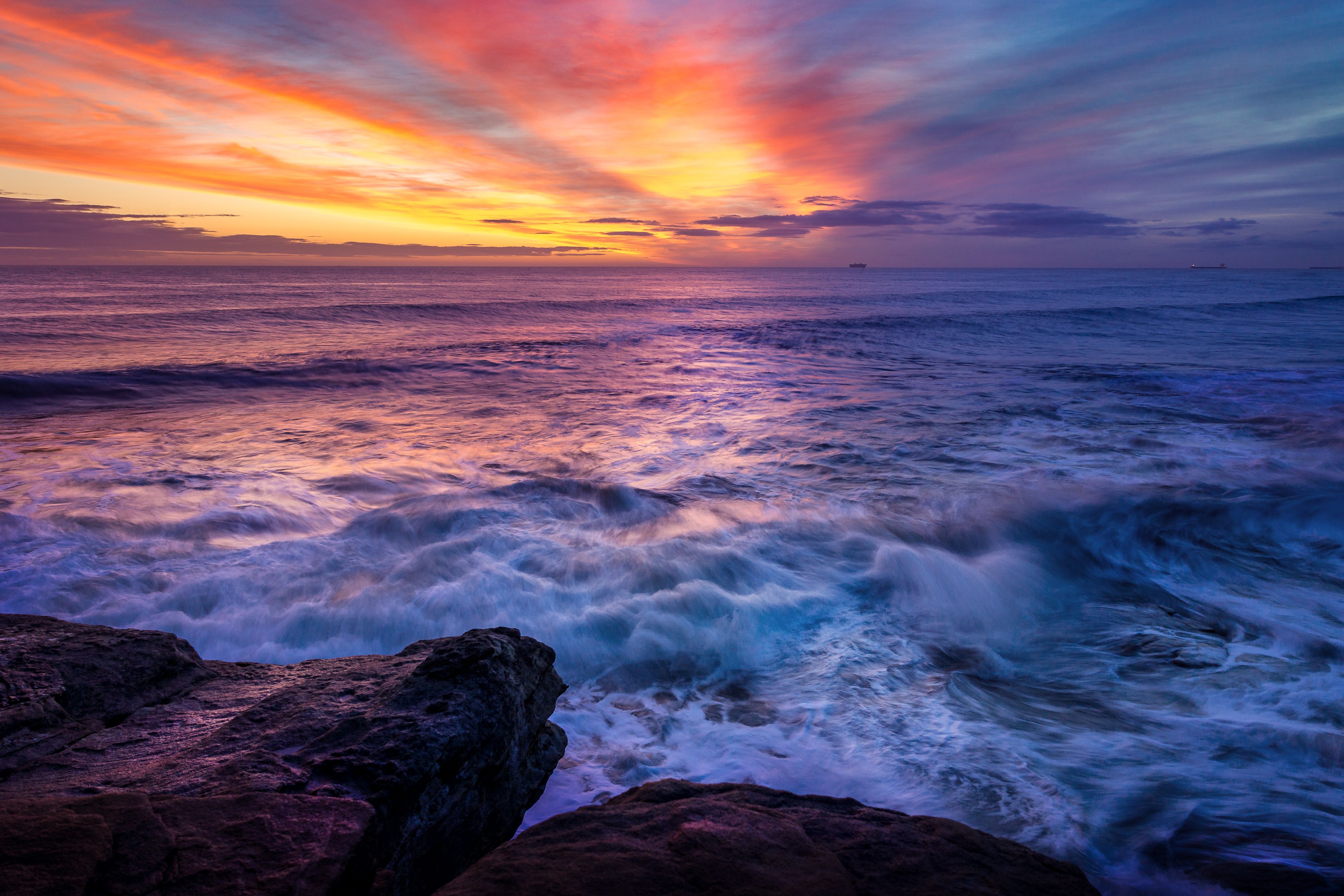 Fotomural Impresionante vista del atardecer sobre las olas del océano rompiendo en la costa rocosa.