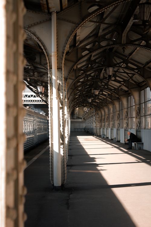 Free A lone person sits under intricate steel arches at an urban railway platform, showing dramatic shadows and light. Stock Photo