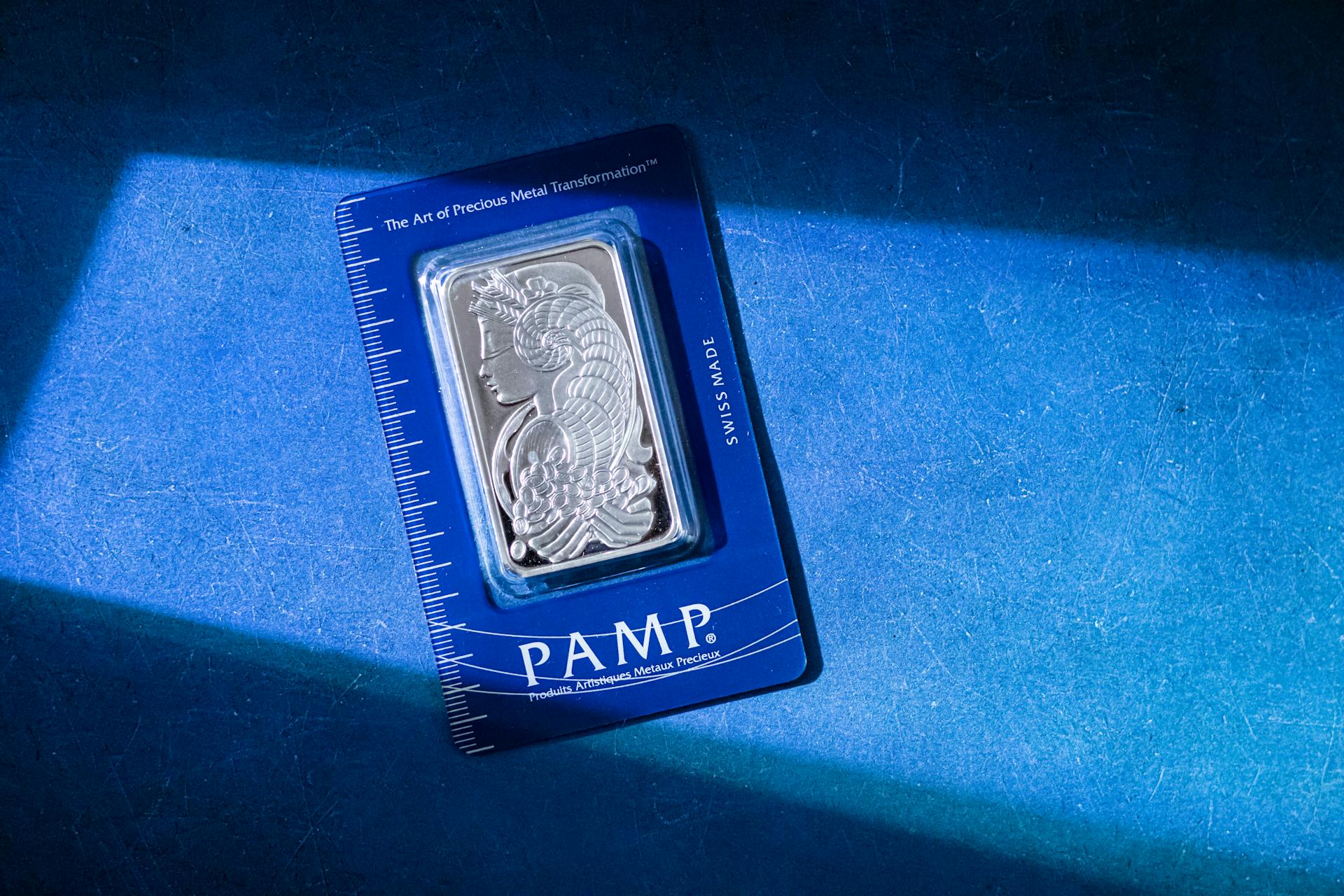 Silver bar in PAMP packaging on a textured blue surface with sunlight.