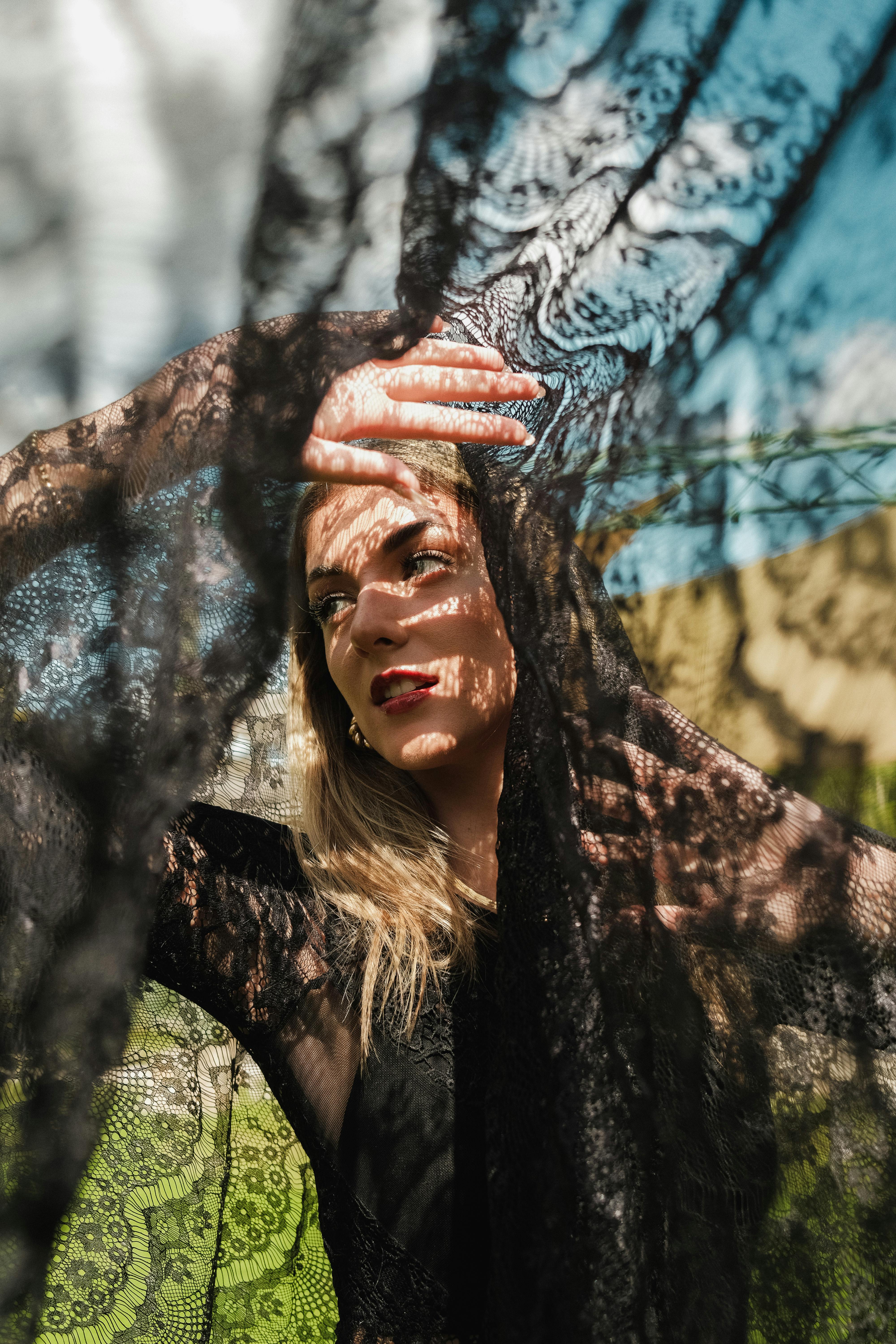 A woman elegantly poses with a black lace veil, creating dramatic shadows under the sunlight.