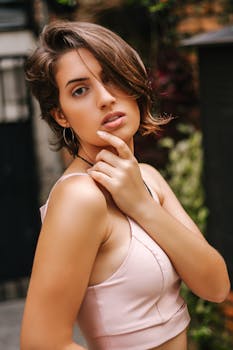 Stylish young woman in a pink top posing confidently outdoors with greenery in the background.