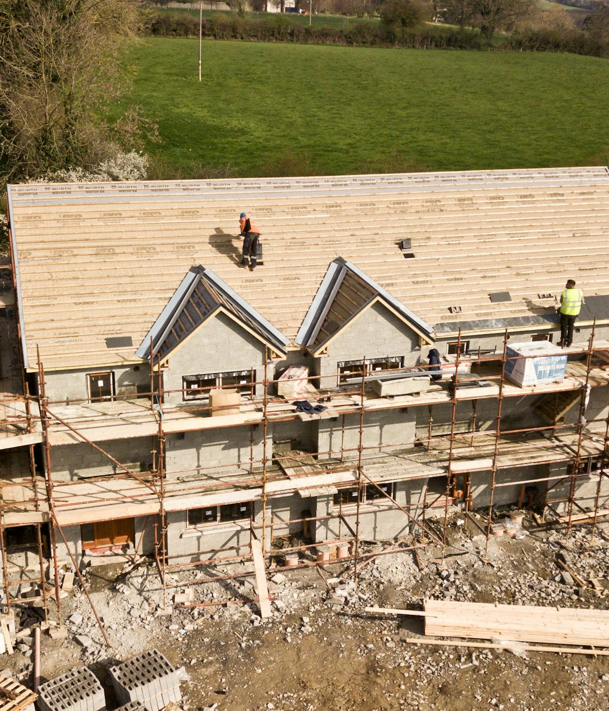 Construction workers on a scaffold are building a house with a wooden roof frame in progress, surrounded by green fields.
