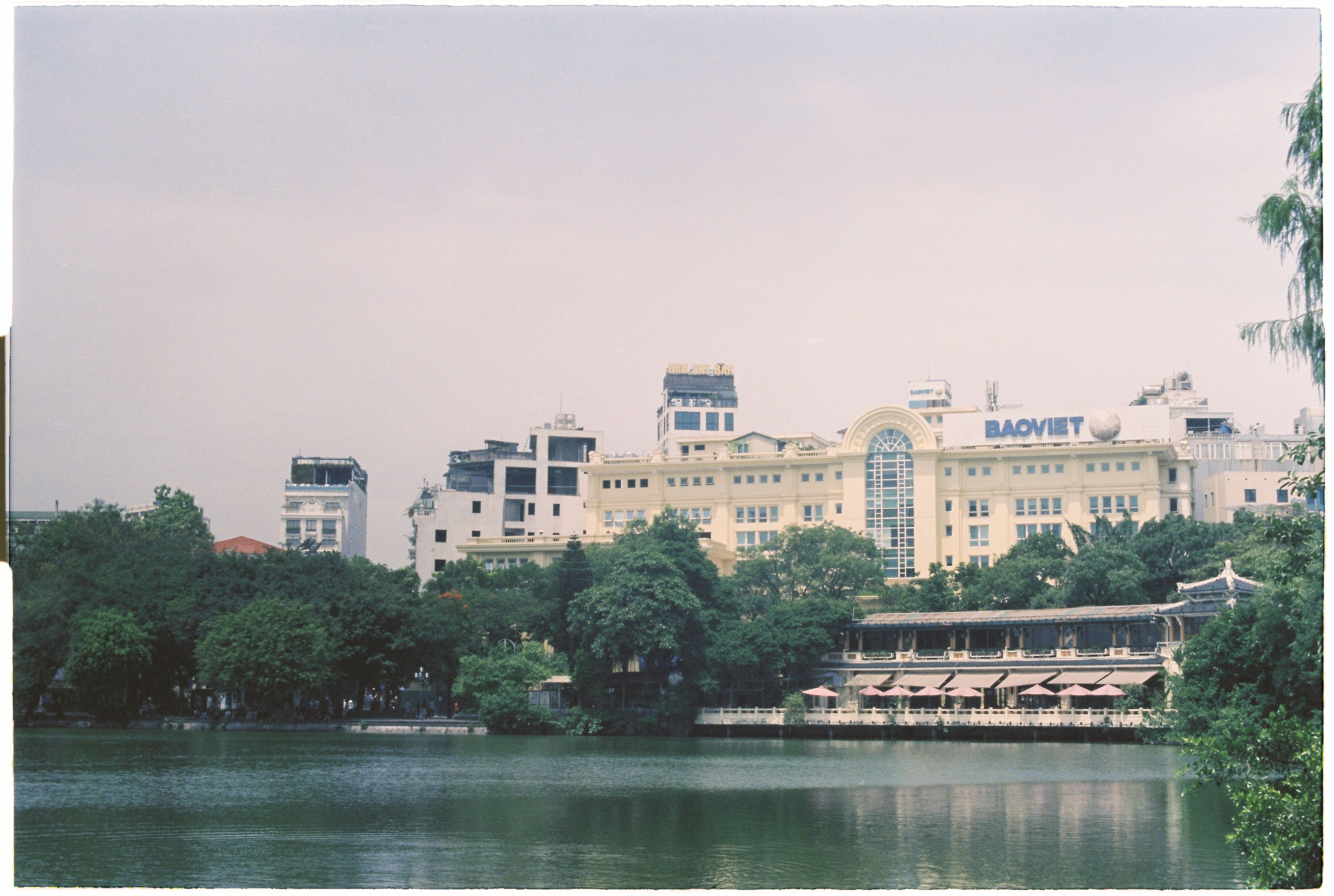 Scenic view of Baoviet building across a lake, framed by trees and urban backdrop.