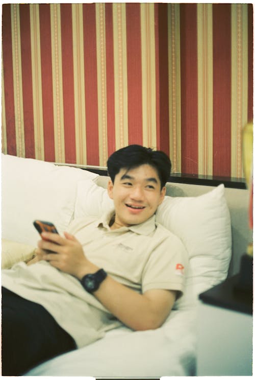 A man smiling while laying on a bed holding a cell phone