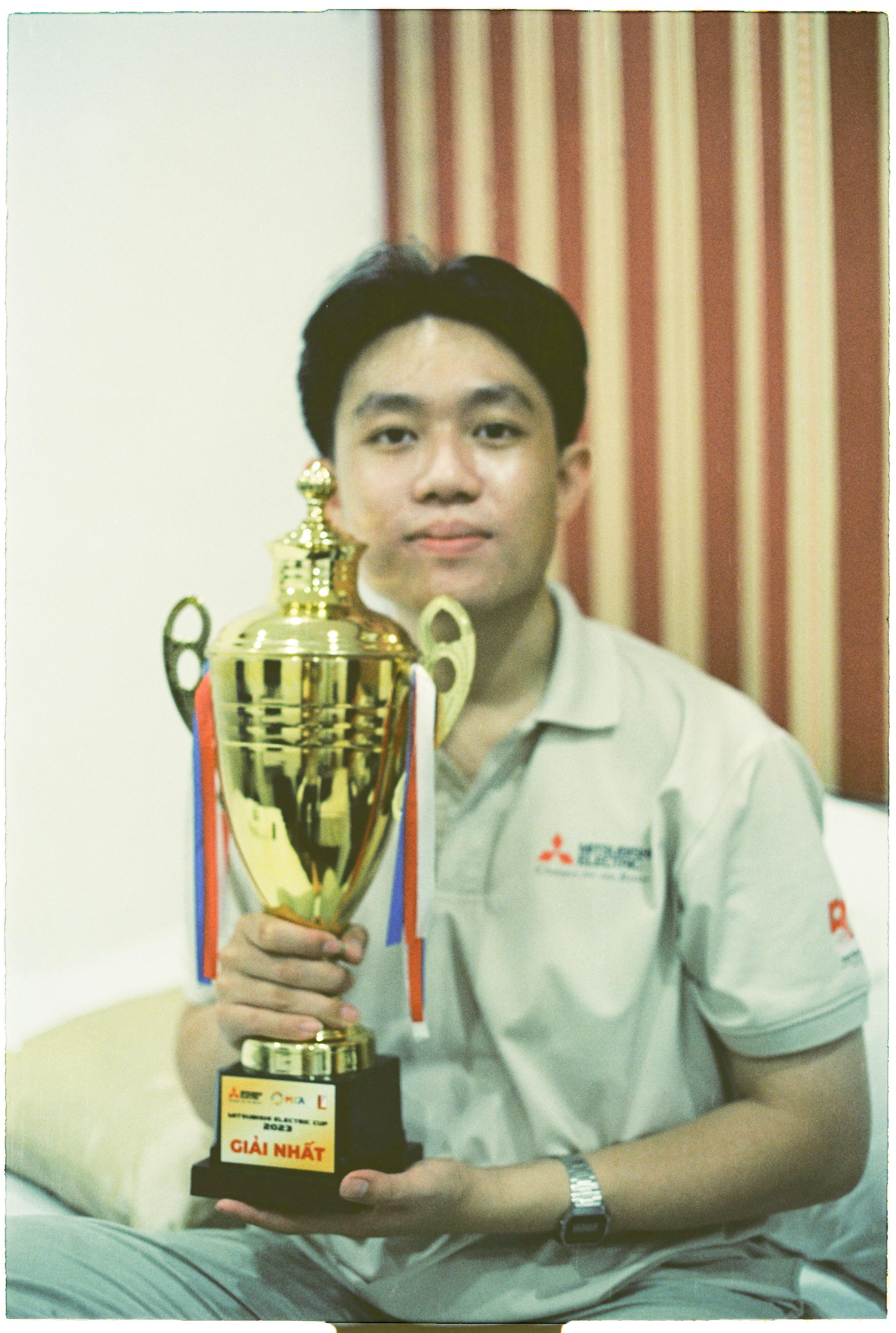 Young Asian man proudly holds a trophy in an indoor setting, representing achievement and success.