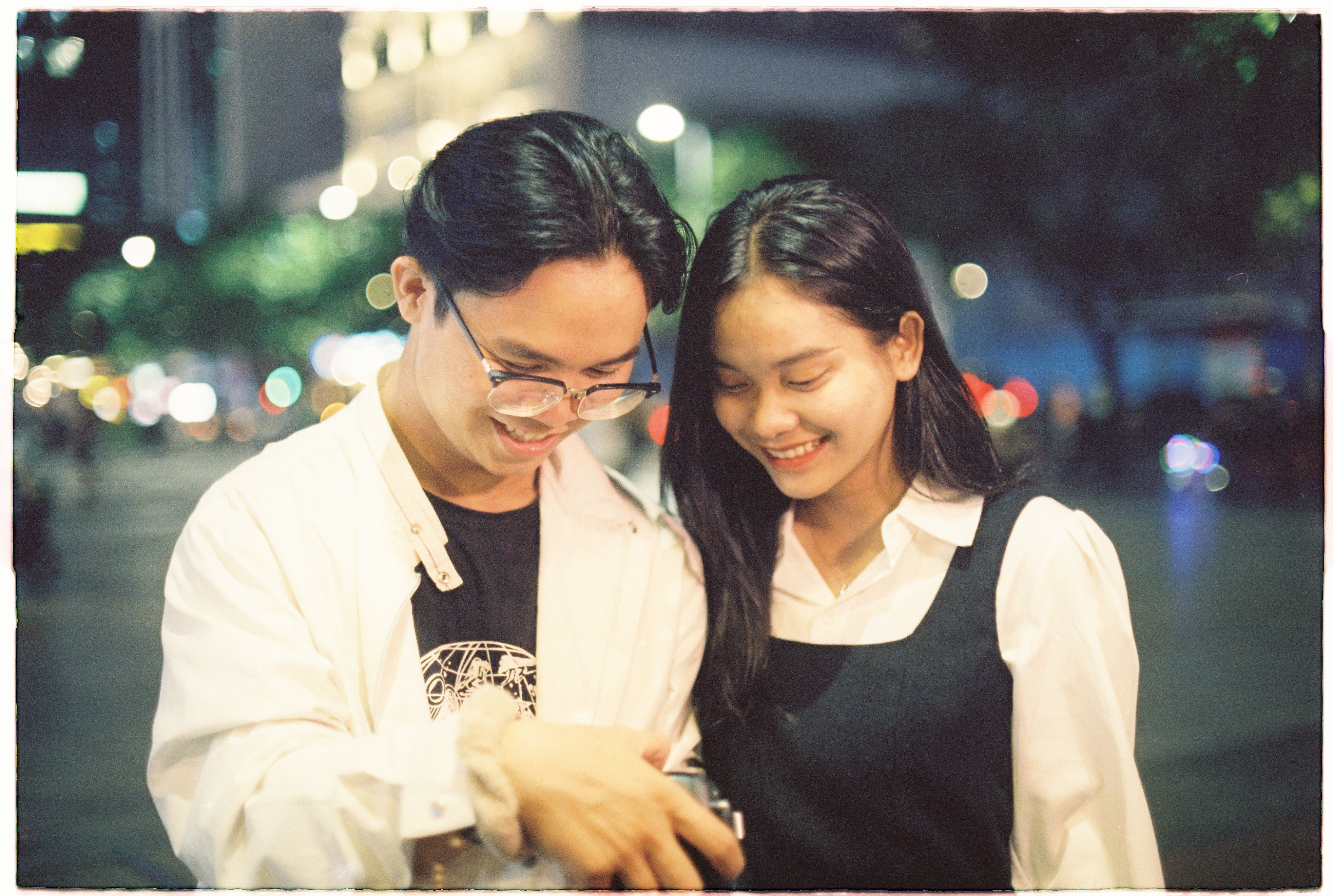 Asian couple smiling and looking down in an urban setting at night, captured in a candid moment.