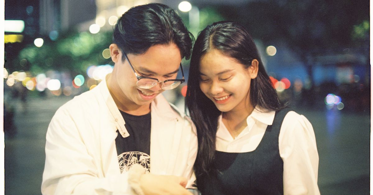 Asian couple smiling and looking down in an urban setting at night, captured in a candid moment.