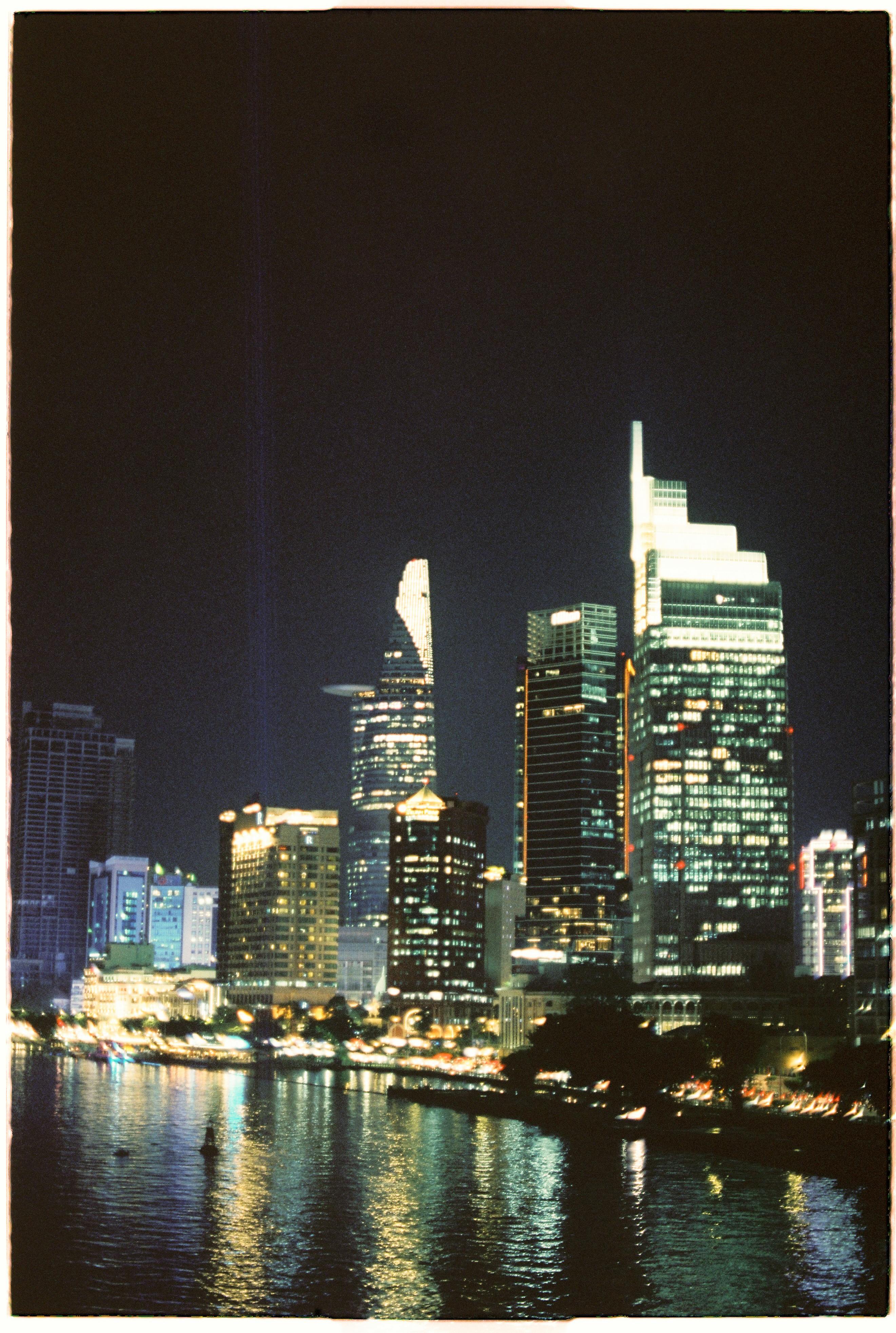 Illuminated Ho Chi Minh City skyline with skyscrapers reflected in the river at night.