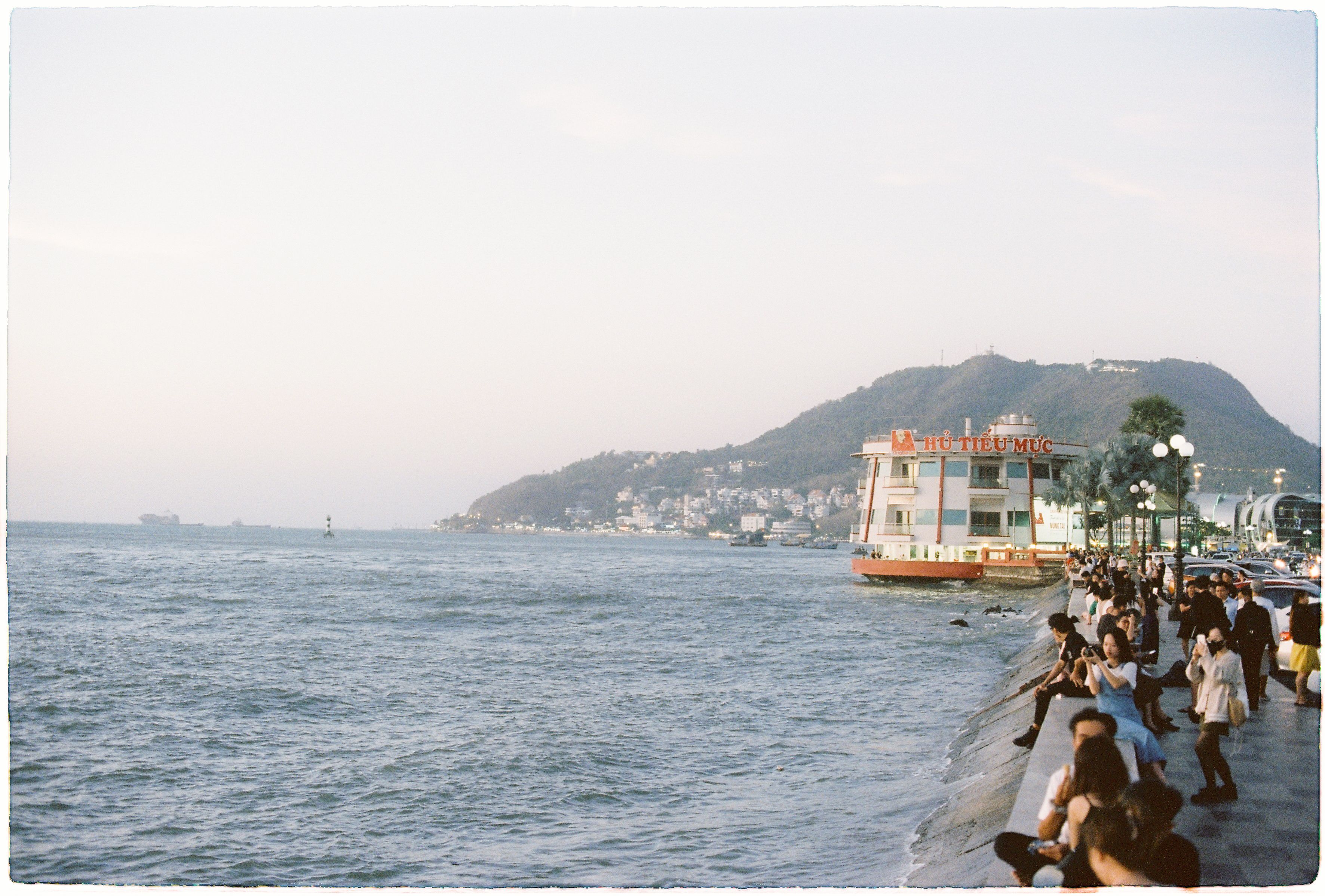 People enjoy a peaceful evening by the waterfront with a view of distant hills and calm seas.