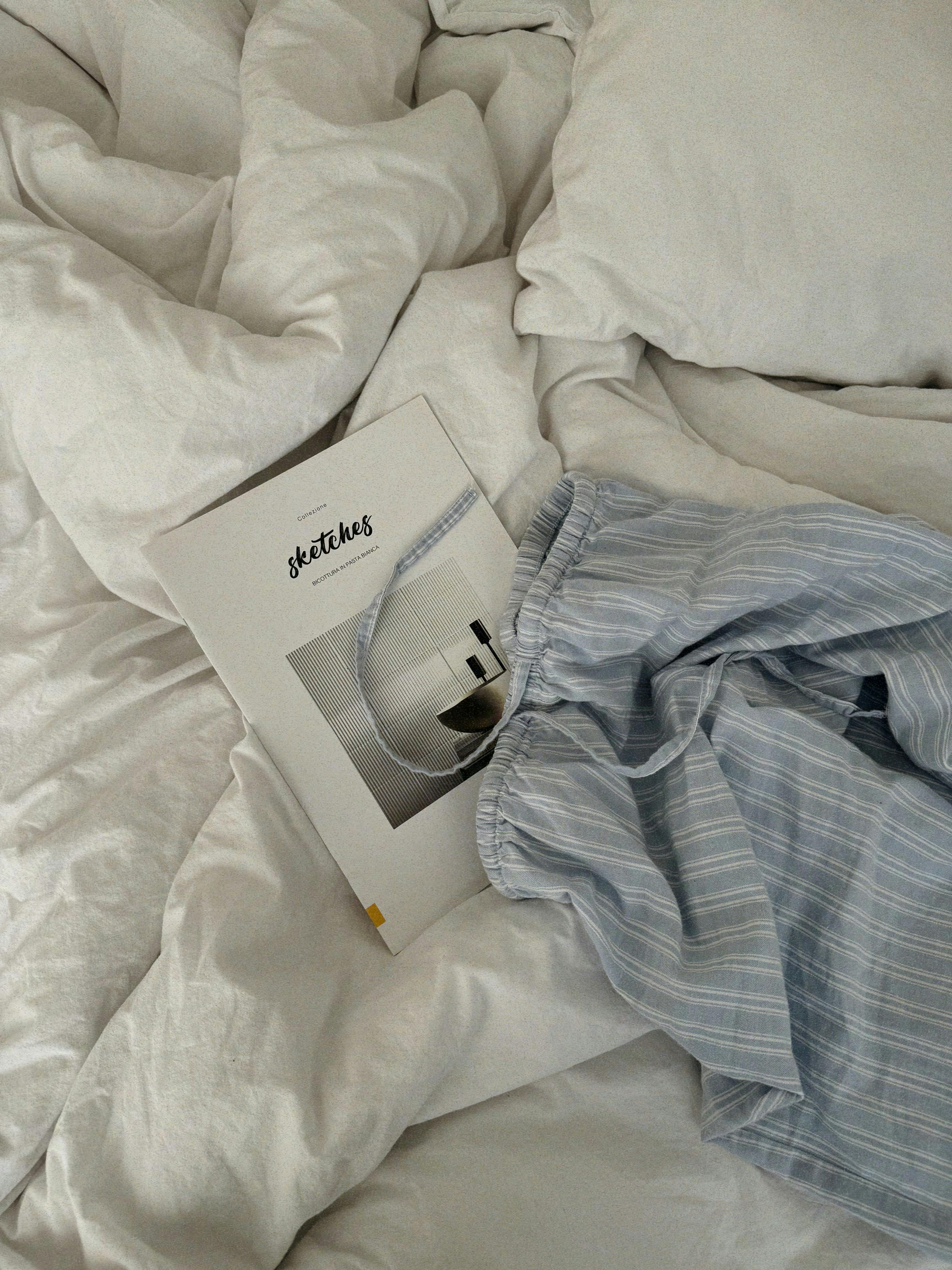 A cozy setup featuring a book and striped pajama pants on a white bed, evoking comfort.