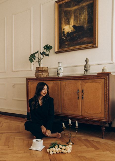A woman in black sits in a stylish vintage room with tulips, a cake, and elegant decor.