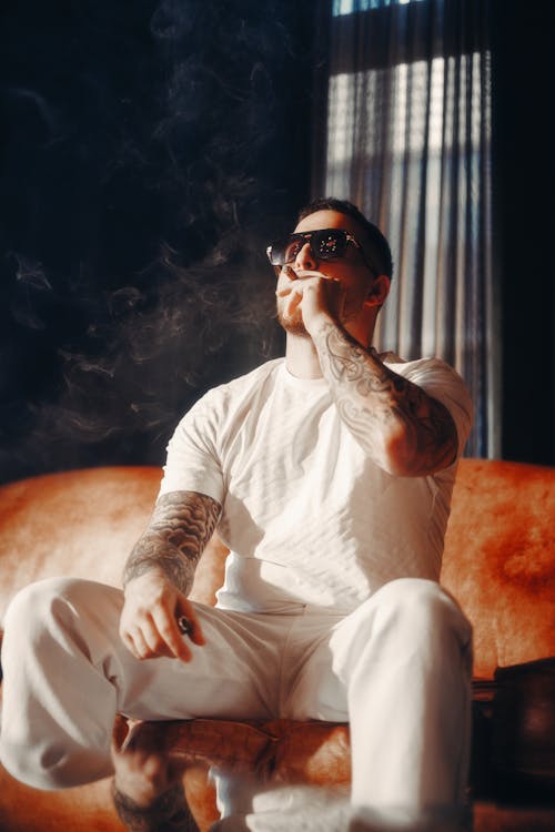 A man in white sitting on a couch smoking a cigarette