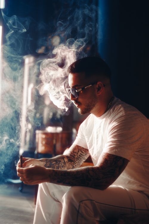 Free Side profile of a tattooed man smoking indoors, enveloped in smoke, wearing sunglasses and a white t-shirt. Stock Photo