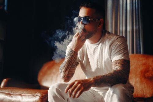 Free Portrait of a tattooed man smoking indoors with sunglasses, surrounded by smoke. Stock Photo