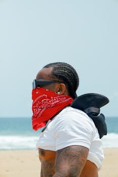 A stylish man with surf gear and a bandana poses at Lagos beach, under a bright sky.