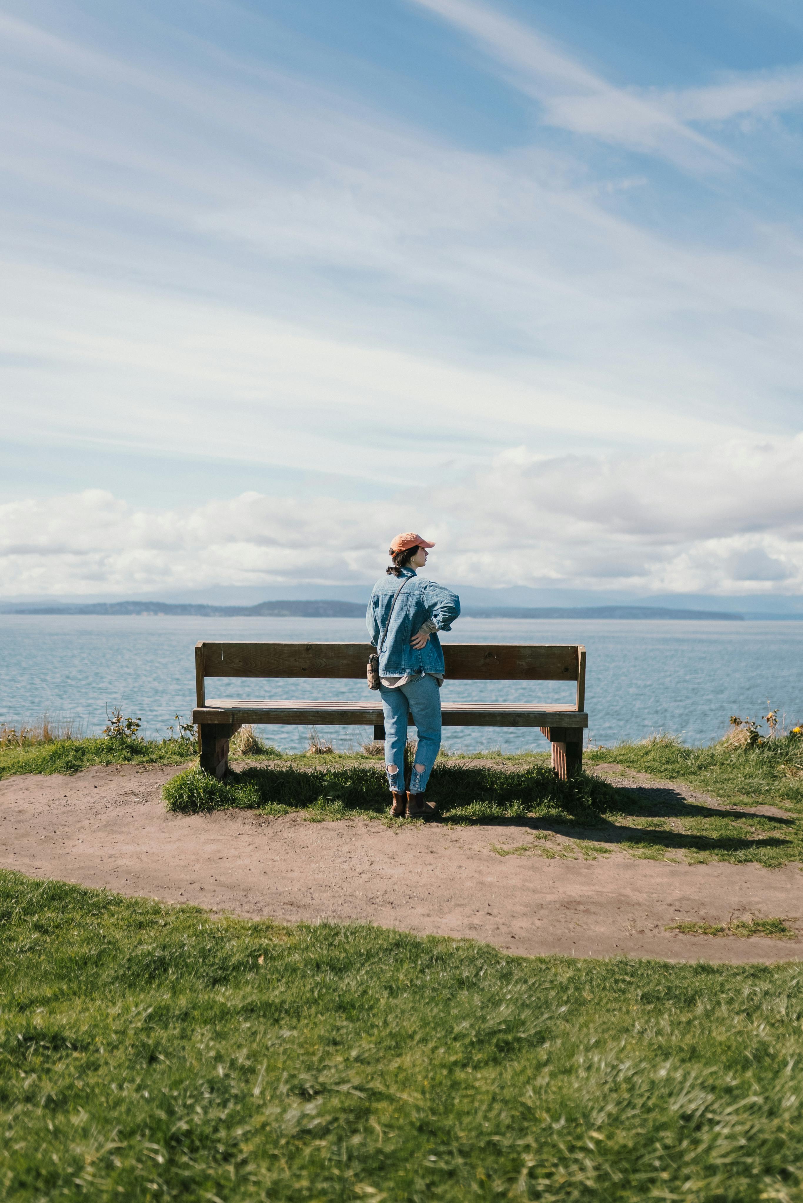 A woman stands by a bench overlooking a serene lakeshore on a sunny day.