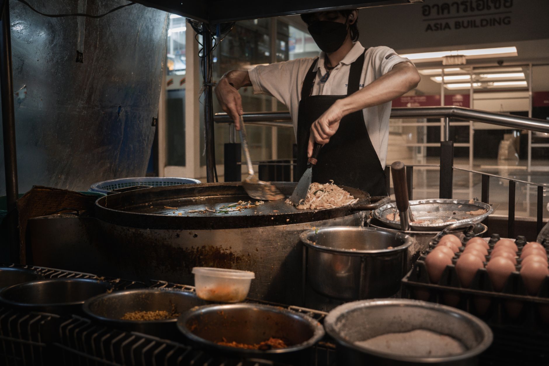 A masked chef skillfully prepares traditional street food in a bustling Bangkok night market.