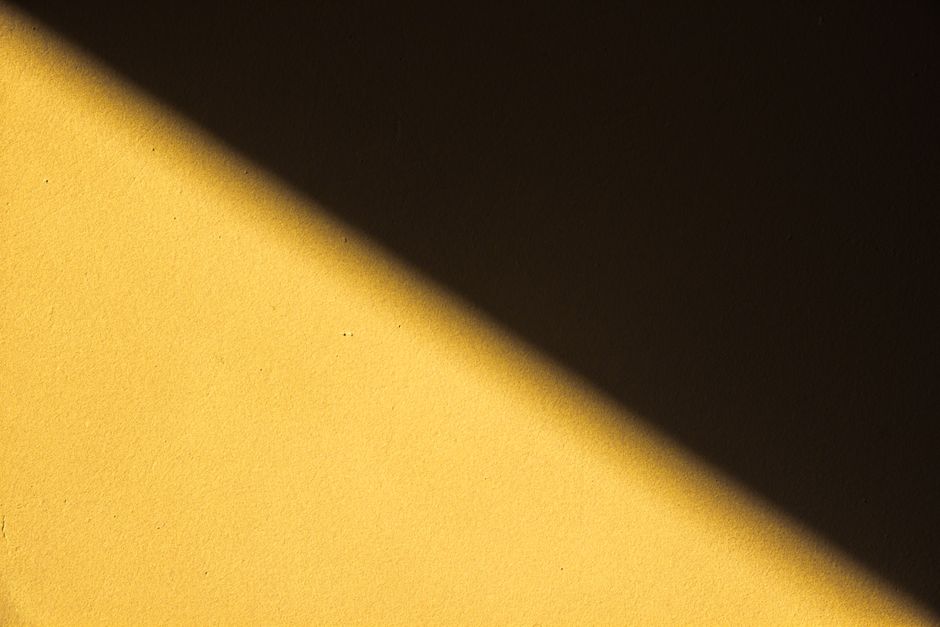 A vivid diagonal shadow contrasts against a textured yellow wall, creating an abstract design.