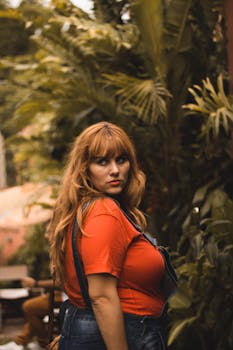 一位身着红色衬衫和牛仔工装裤的女性时尚肖像，被郁郁葱葱的绿色植物所环绕