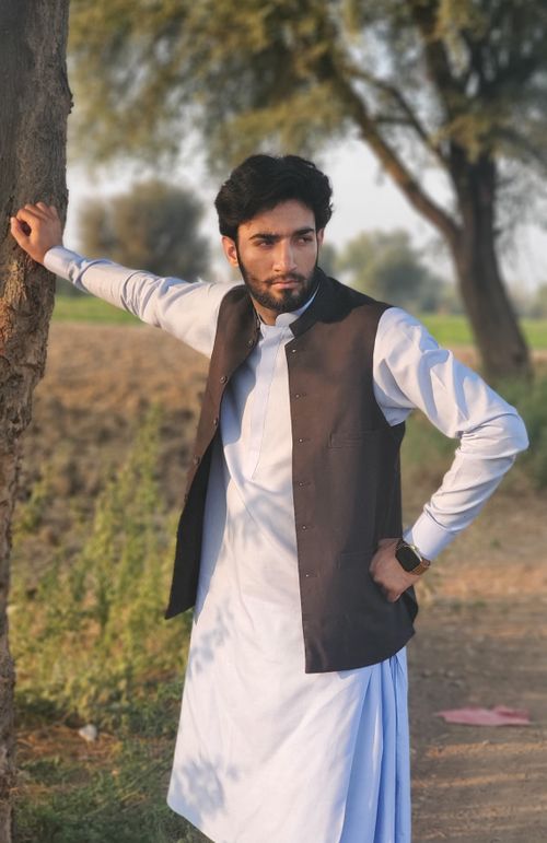 Free A man stands outdoors in traditional attire, leaning against a tree with a thoughtful expression in a natural setting. Stock Photo